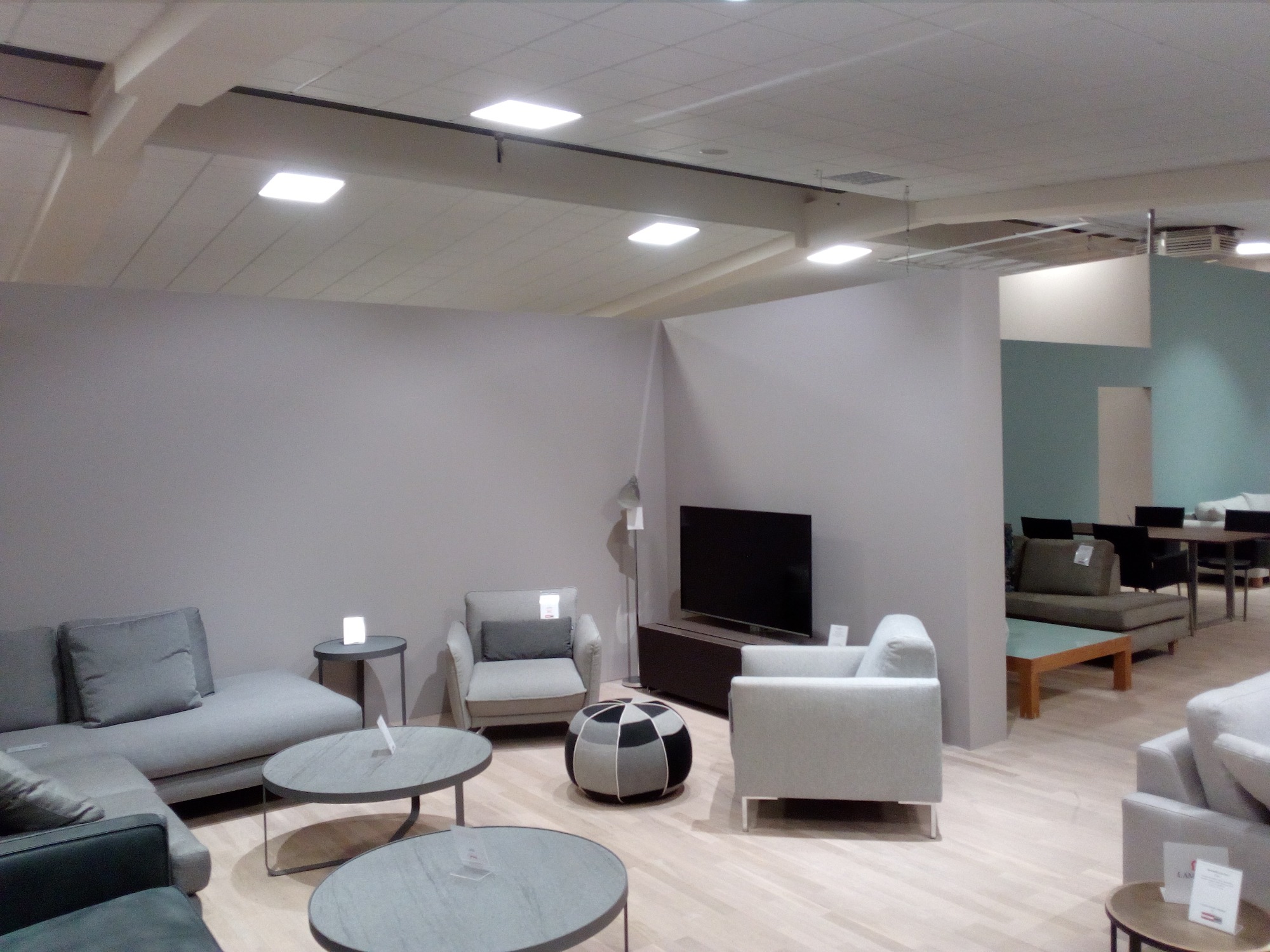 Jasne, przestronne wnętrze z szarą sofą narożną, fotelami i okrągłymi stolikami kawowymi na drewnianej podłodze, oświetlone kwadratowymi lampami sufitowymi.