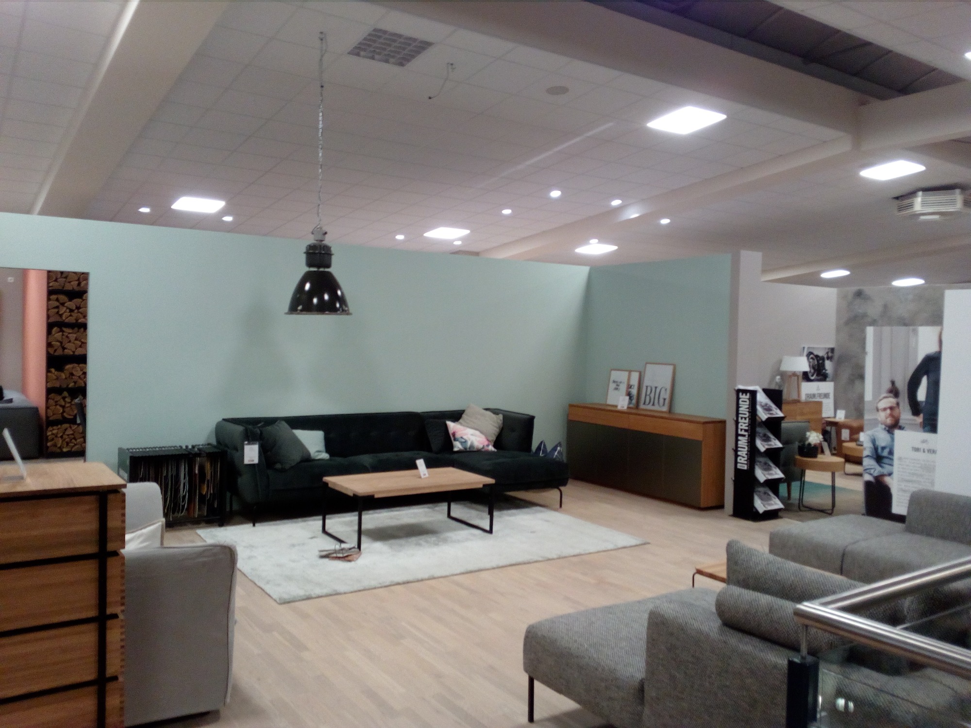 Jasne, przestronne wnętrze ekspozycyjne z sofą, stolikiem kawowym i lampą wiszącą w stylu industrialnym, ściany pomalowane na jasny, pastelowy kolor.