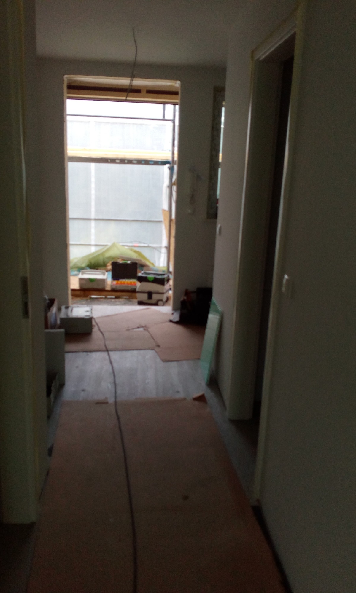 Korytarz w trakcie remontu: podłoga zabezpieczona kartonem, widoczne narzędzia i materiały budowlane w oddali, okno z widocznym rusztowaniem na zewnątrz.