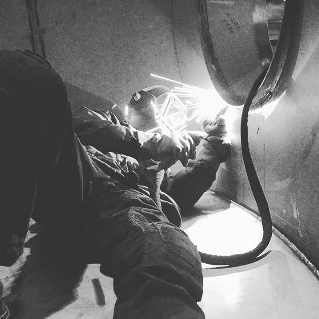 Czarno-białe zdjęcie spawacza w hełmie ochronnym, pracującego wewnątrz metalowej konstrukcji, widoczne iskry i kabel spawalniczy.