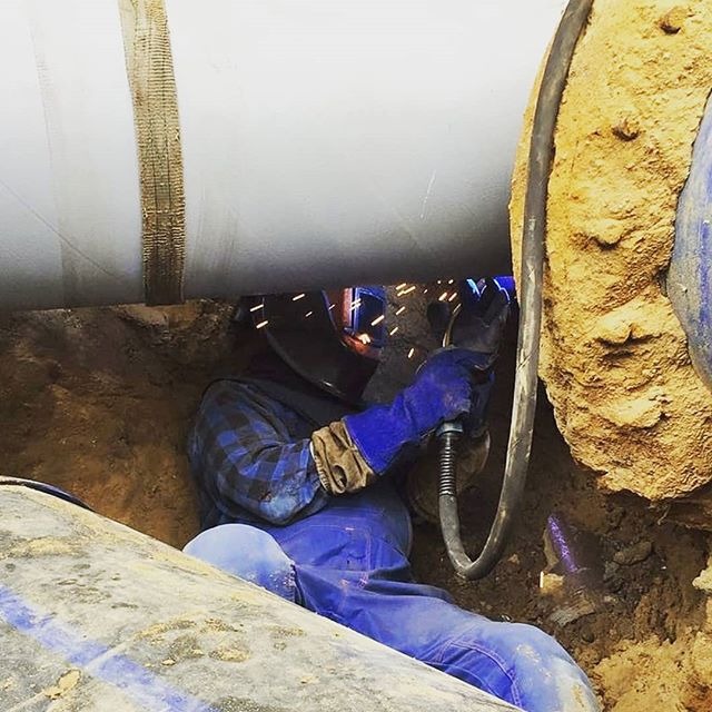 Pracownik w niebieskim kombinezonie i masce spawalniczej łączy rury w wykopie, iskry podczas spawania, widoczne złącze i kabel spawalniczy.