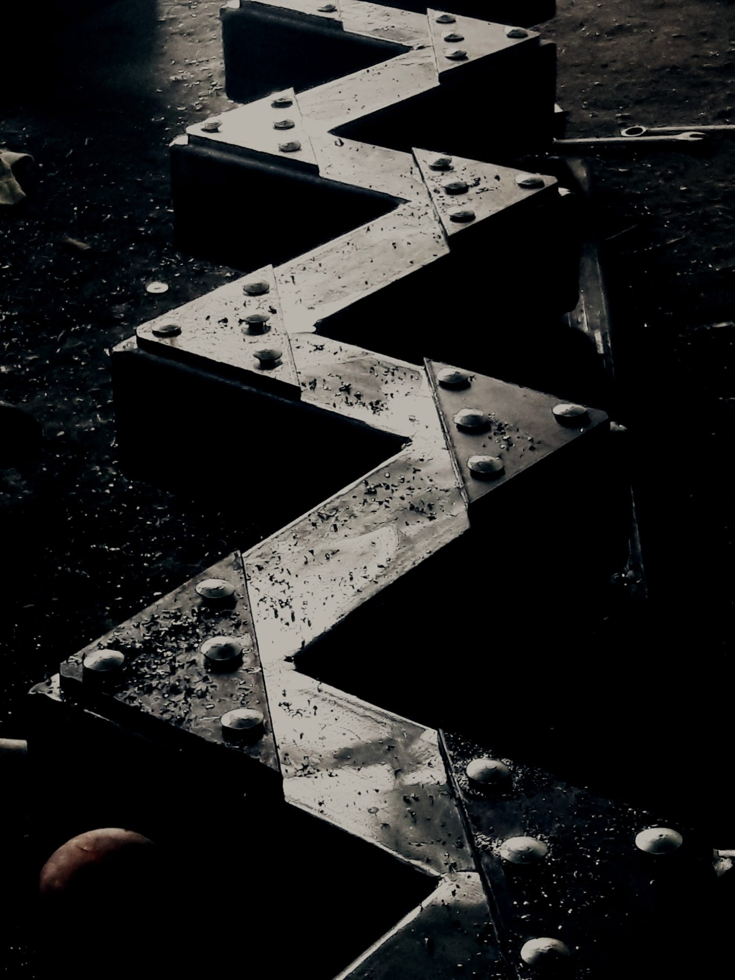 Metalowa konstrukcja w kształcie zygzaka, łączona nitami, z widocznymi śladami korozji i narzędziami w tle, w ciemnym otoczeniu.