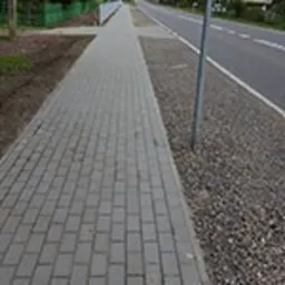 Szary chodnik z kostki brukowej ułożony wzdłuż asfaltowej drogi, oddzielony pasem żwiru. Widok z perspektywy pieszego, w tle słup drogowy i zieleń.