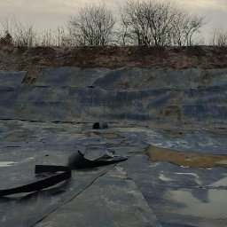 P-MONT PAWEŁ PŁOCIŃSKI - Wykładanie folią dna wykopu pod zbiornik wodny, widoczne fragmenty folii, rury i taśmy mocującej, w tle ziemny brzeg wykopu z rzadką roślinnością, pochmurne niebo.