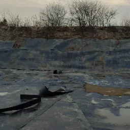 Wykładanie folią dna wykopu pod zbiornik wodny, widoczne fragmenty folii, rury i taśmy mocującej, w tle ziemny brzeg wykopu z rzadką roślinnością, pochmurne niebo.