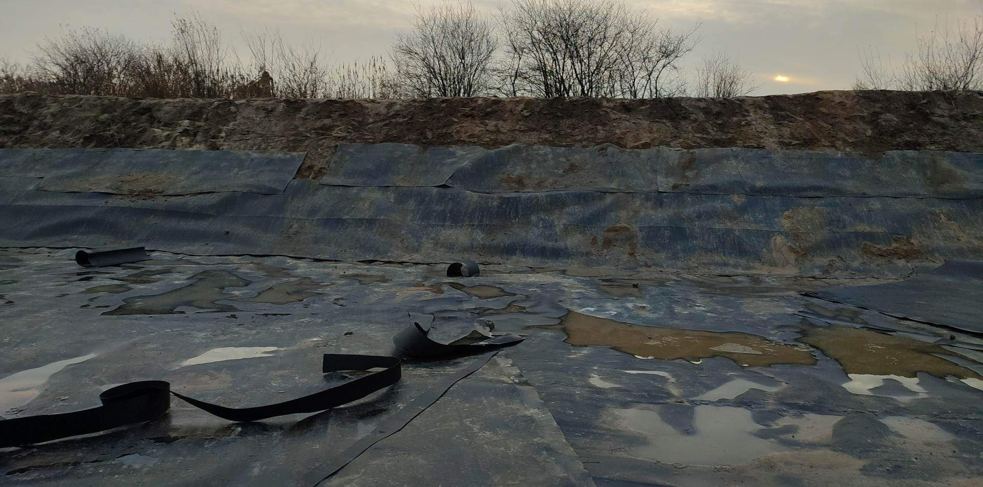 Wykładanie folią dna wykopu pod zbiornik wodny, widoczne fragmenty folii, rury i taśmy mocującej, w tle ziemny brzeg wykopu z rzadką roślinnością, pochmurne niebo.