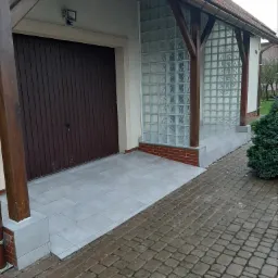 Wyłożona szarymi płytkami podłoga przed garażem z drewnianymi wrotami i ścianą z luksferów, widok na kostkę brukową.