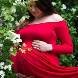 Agnieszka Szpak FOTOGRAFIA - Kobieta w czerwonej sukni ciążowej pozuje na tle kwitnącego krzewu z białymi kwiatami, dłonie spoczywają na brzuchu, delikatny uśmiech.