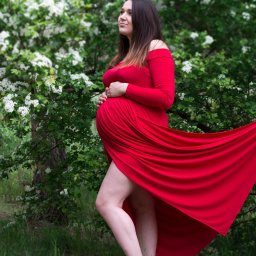 Agnieszka Szpak FOTOGRAFIA - Kobieta w czerwonej sukni ciążowej, stojąca boso na trawie przed kwitnącym krzewem, z rękami na brzuchu i wzrokiem skierowanym w górę.