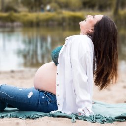 Agnieszka Szpak FOTOGRAFIA - Uśmiechnięta, przyszła mama w jeansach z dziurami i rozpiętej białej koszuli, siedząca na plaży na tle jeziora, w trakcie sesji zdjęciowej.
