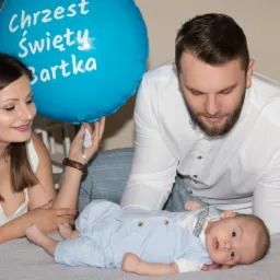 Rodzice spoglądają na leżącego na szarym kocu niemowlaka ubranego w błękitny kombinezon, obok błękitny balon z napisem 'Chrzest Święty Bartka'