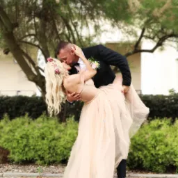 Para młoda w tańcu, panna młoda w jasnej sukni i wianku z kwiatów, pan młody w czarnym garniturze, pocałunek na tle zieleni.