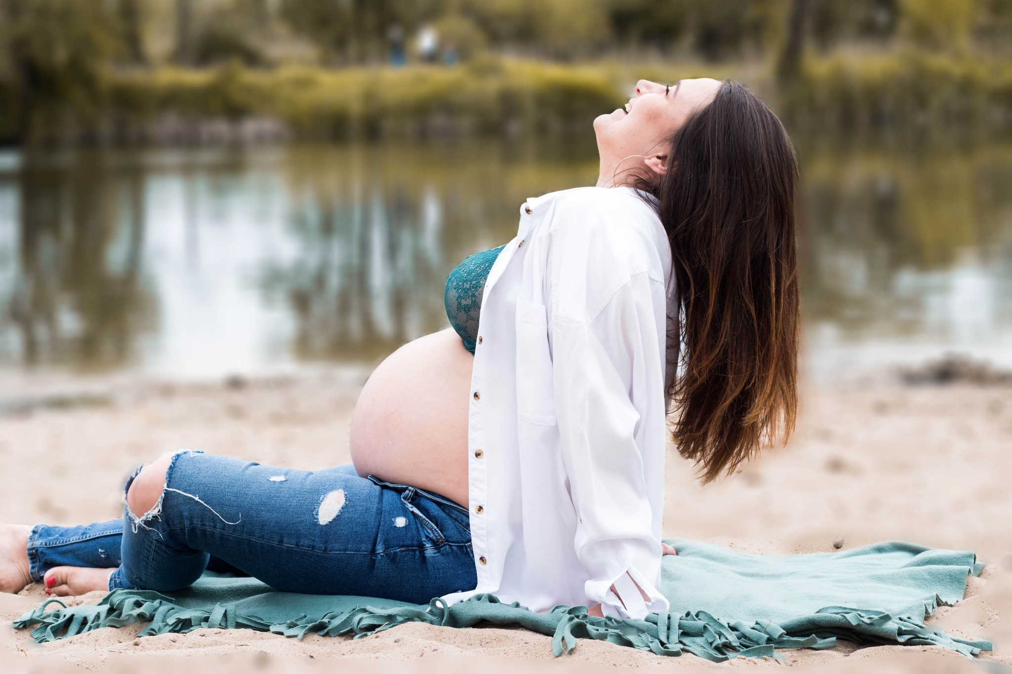 Uśmiechnięta, przyszła mama w jeansach z dziurami i rozpiętej białej koszuli, siedząca na plaży na tle jeziora, w trakcie sesji zdjęciowej.