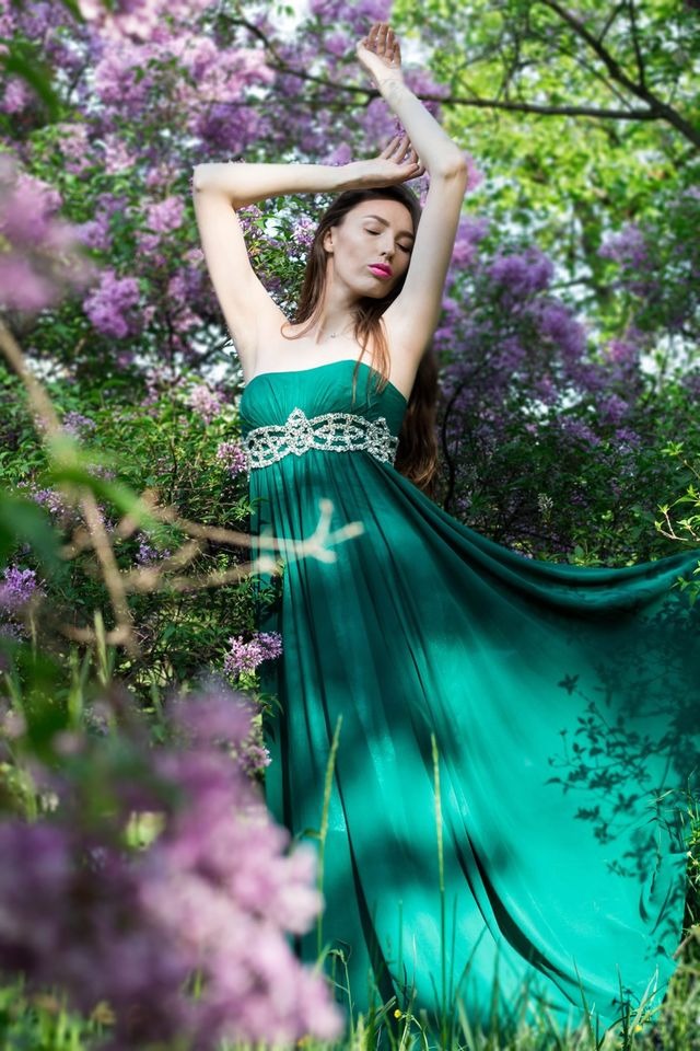 Kobieta w długiej, szmaragdowej sukni bez ramiączek z ozdobnym pasem, pozująca z uniesionymi rękami na tle kwitnącego bzu w słoneczny dzień.