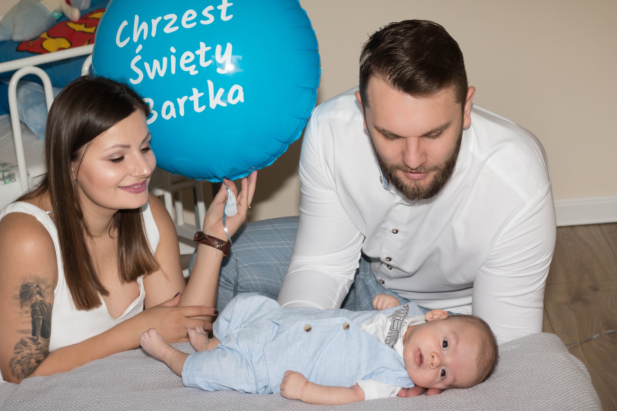 Rodzice spoglądają na leżącego na szarym kocu niemowlaka ubranego w błękitny kombinezon, obok błękitny balon z napisem 'Chrzest Święty Bartka'