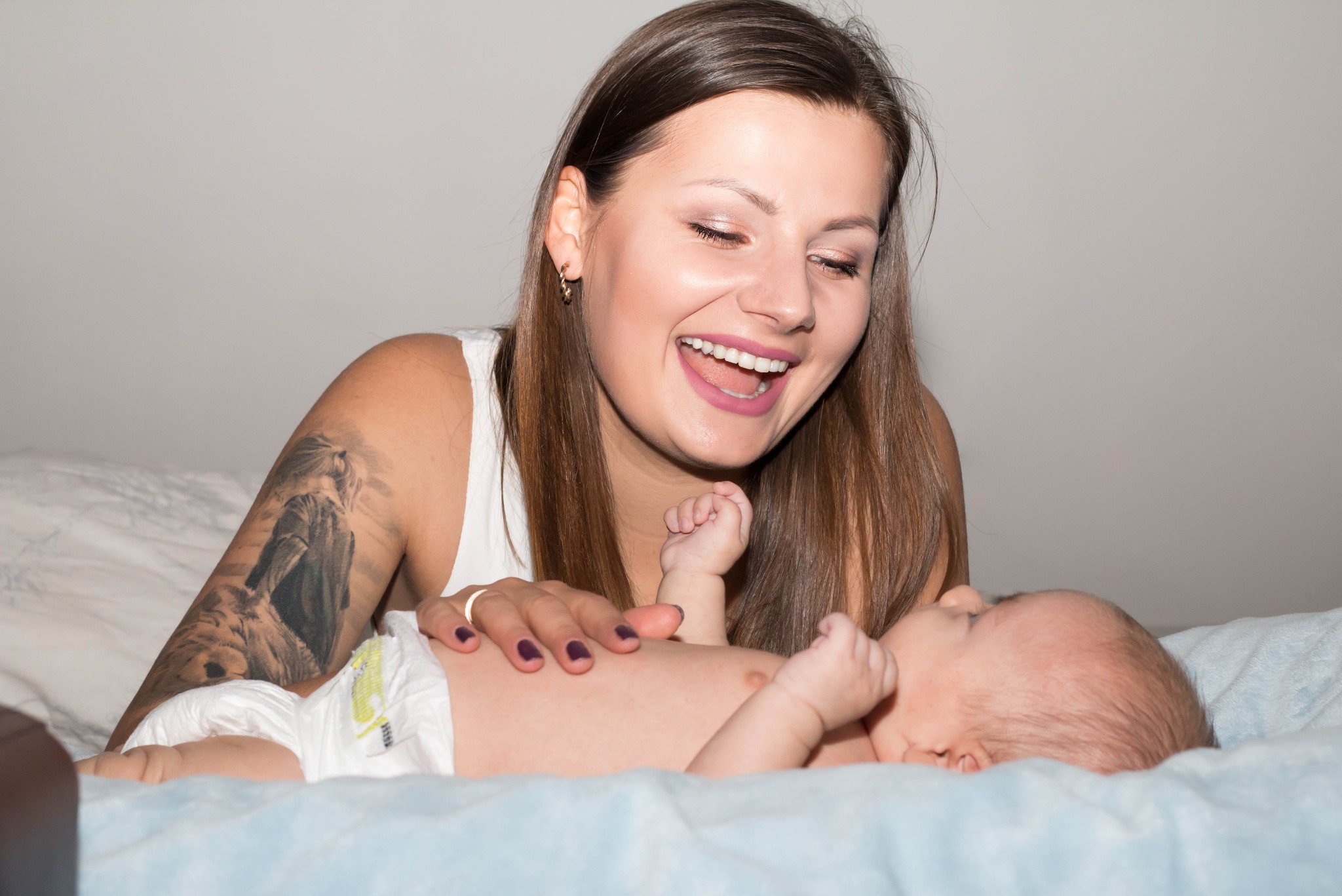 Uśmiechnięta kobieta z tatuażem na ramieniu leży na łóżku z niemowlęciem w pieluszce.