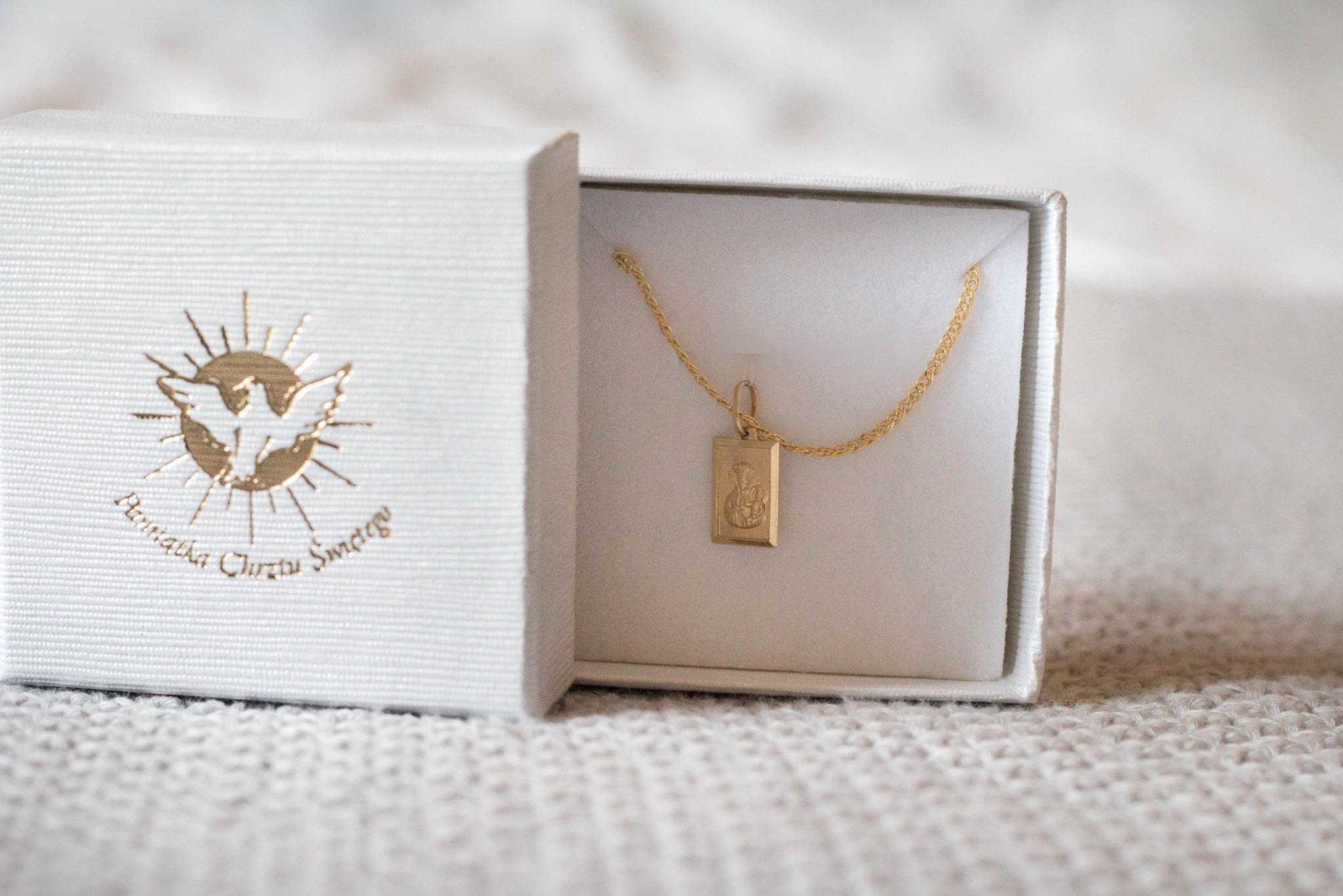 Złoty łańcuszek z medalikiem w białym pudełku z napisem 'Pamiątka Chrztu Świętego' i symbolem gołębicy na beżowym tle.