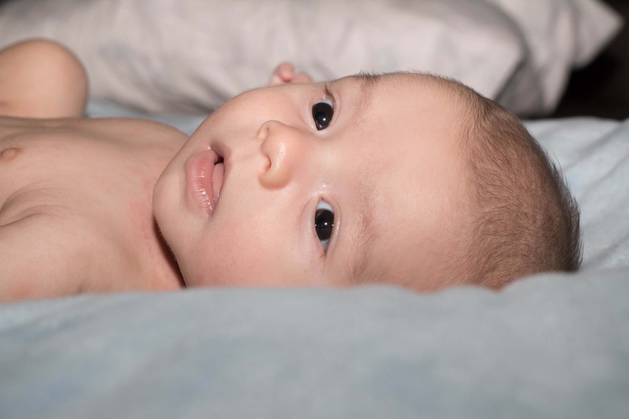 Noworodek leżący na plecach, z otwartymi, ciemnymi oczami i lekko otwartymi ustami, na jasnoniebieskim tle.
