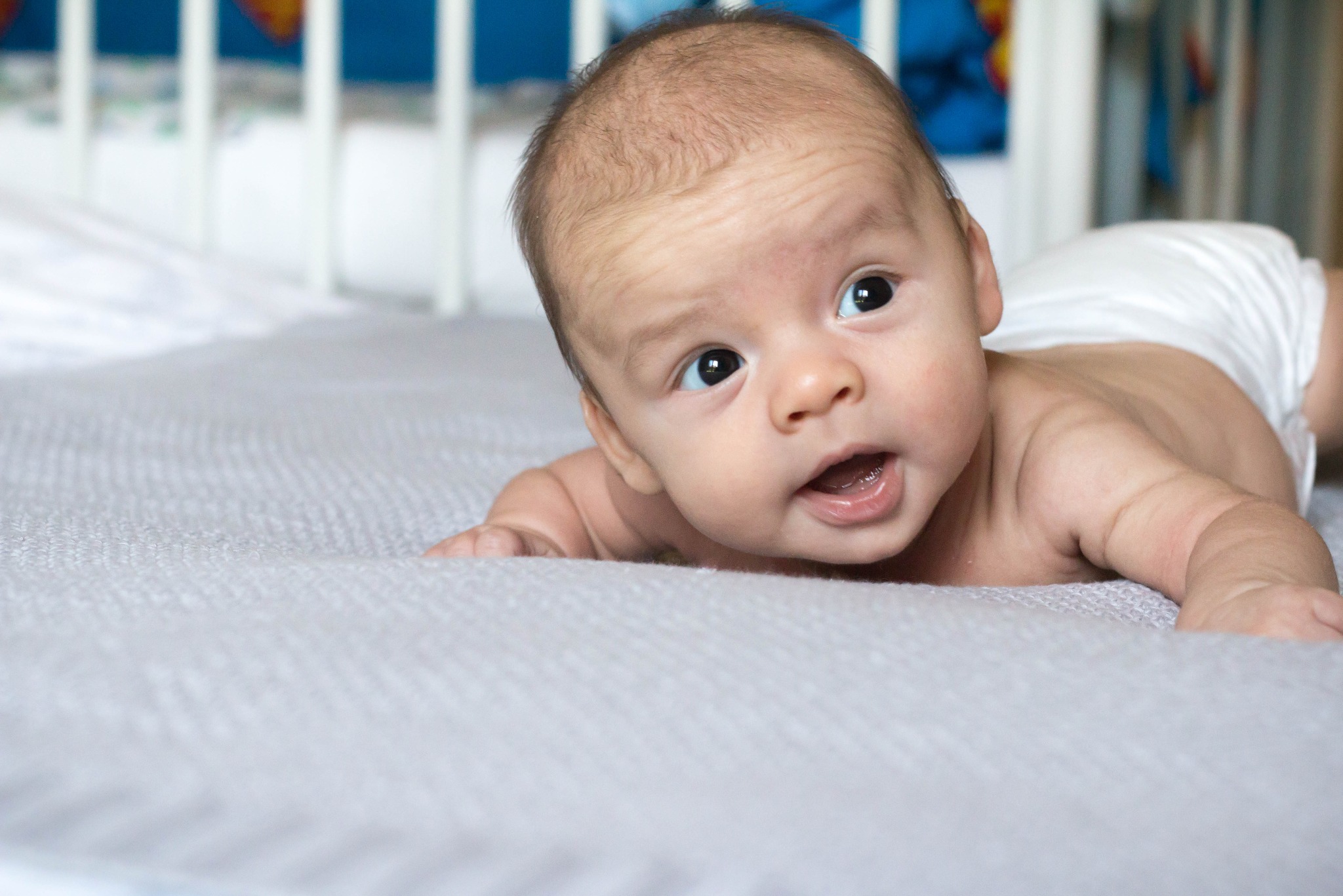 Noworodek leży na brzuchu na szarym kocu, z otwartymi ustami i zaciekawionym spojrzeniem, pieluszka na pupie, w tle białe łóżeczko.