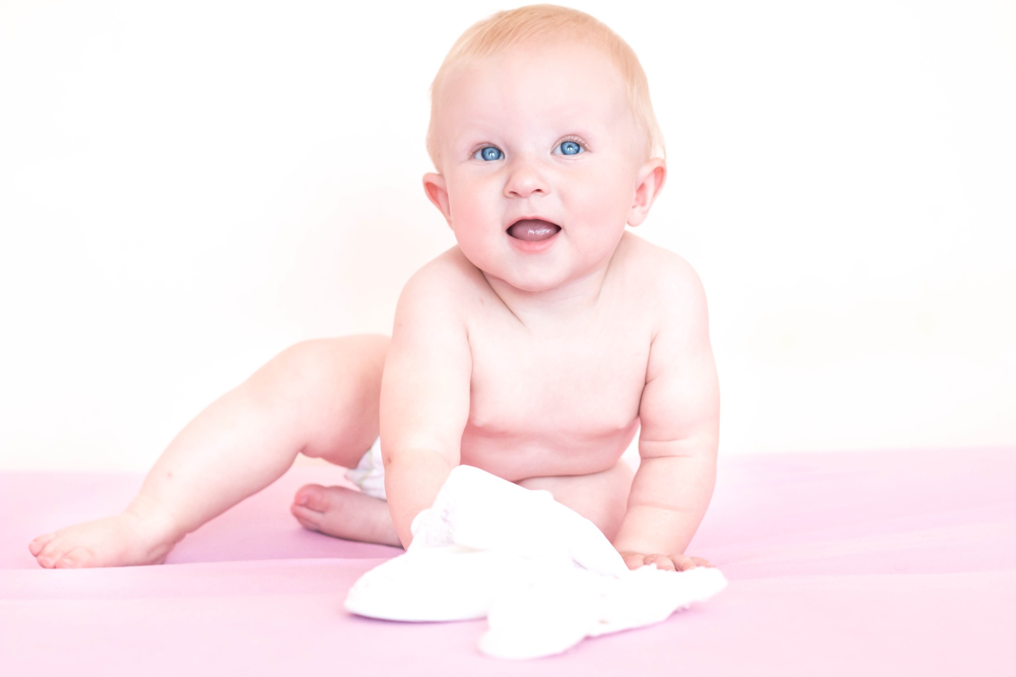 Uśmiechnięty, jasnowłosy niemowlak z niebieskimi oczami, leżący na brzuchu na różowym podłożu, trzymający w dłoni białą pieluszkę tetrową.
