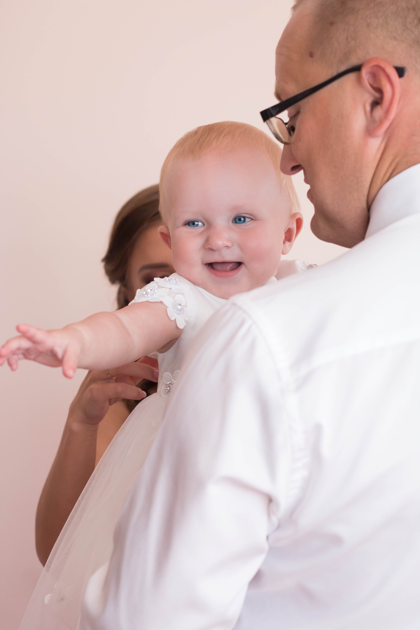 Uśmiechnięta niemowlęca dziewczynka w białej sukience z kwiatowymi aplikacjami, trzymana na rękach przez mężczyznę w okularach, z matką w tle.