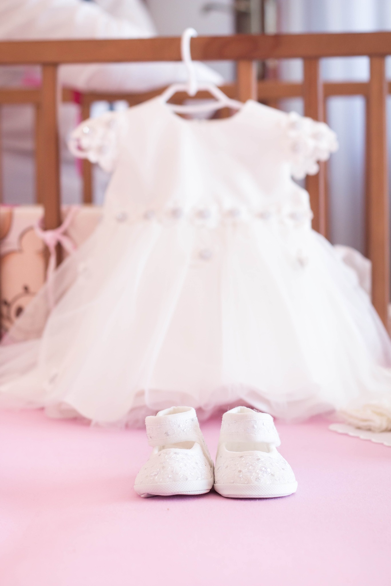 Biała sukienka niemowlęca z aplikacjami kwiatowymi zawieszona na wieszaku nad parą białych, zdobionych bucików na różowym tle.