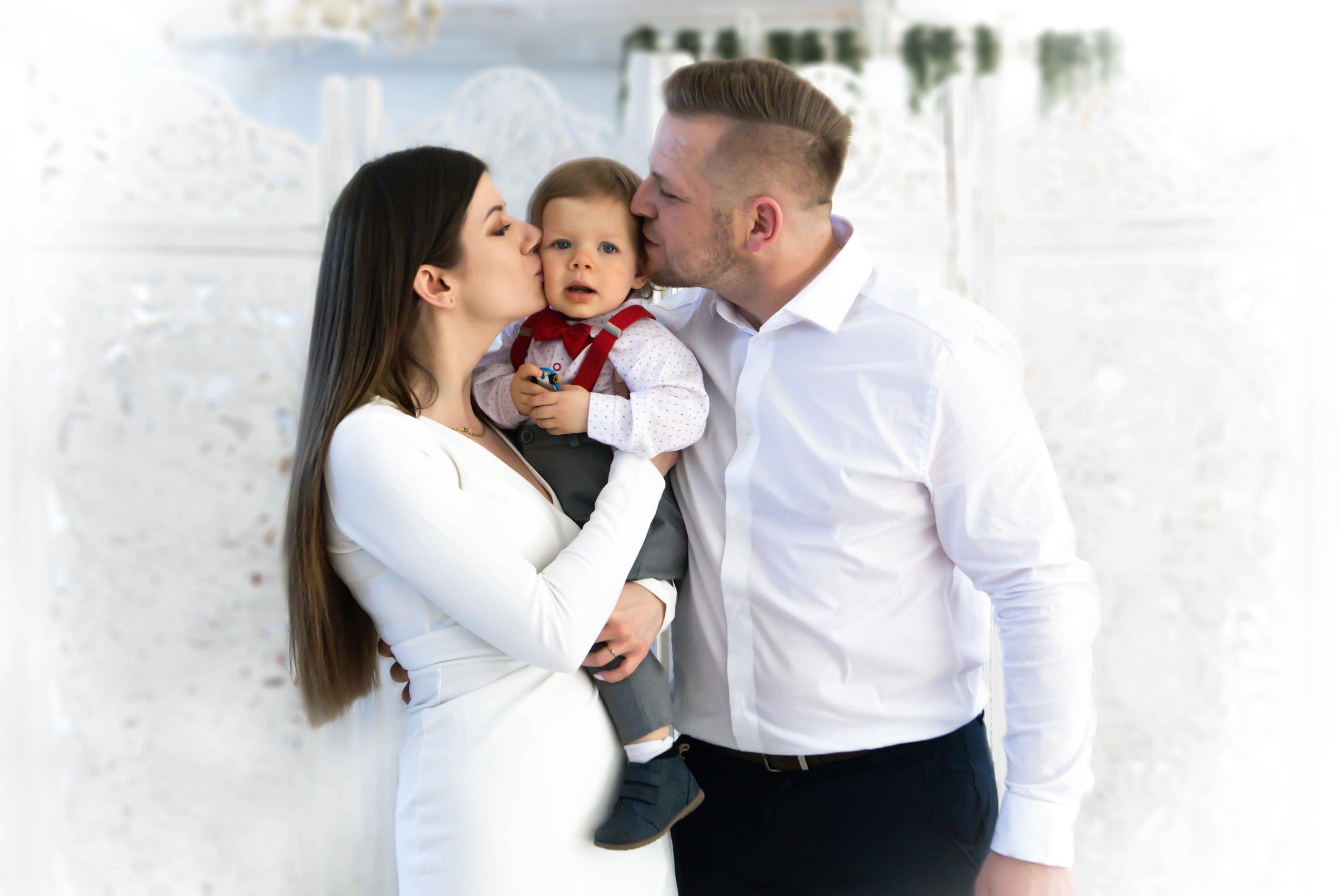 Portret rodzinny: mama w białej sukience i tata w białej koszuli całują w policzki rocznego synka w szarym garniturze i czerwonej muszce na tle białej dekoracyjnej ściany.