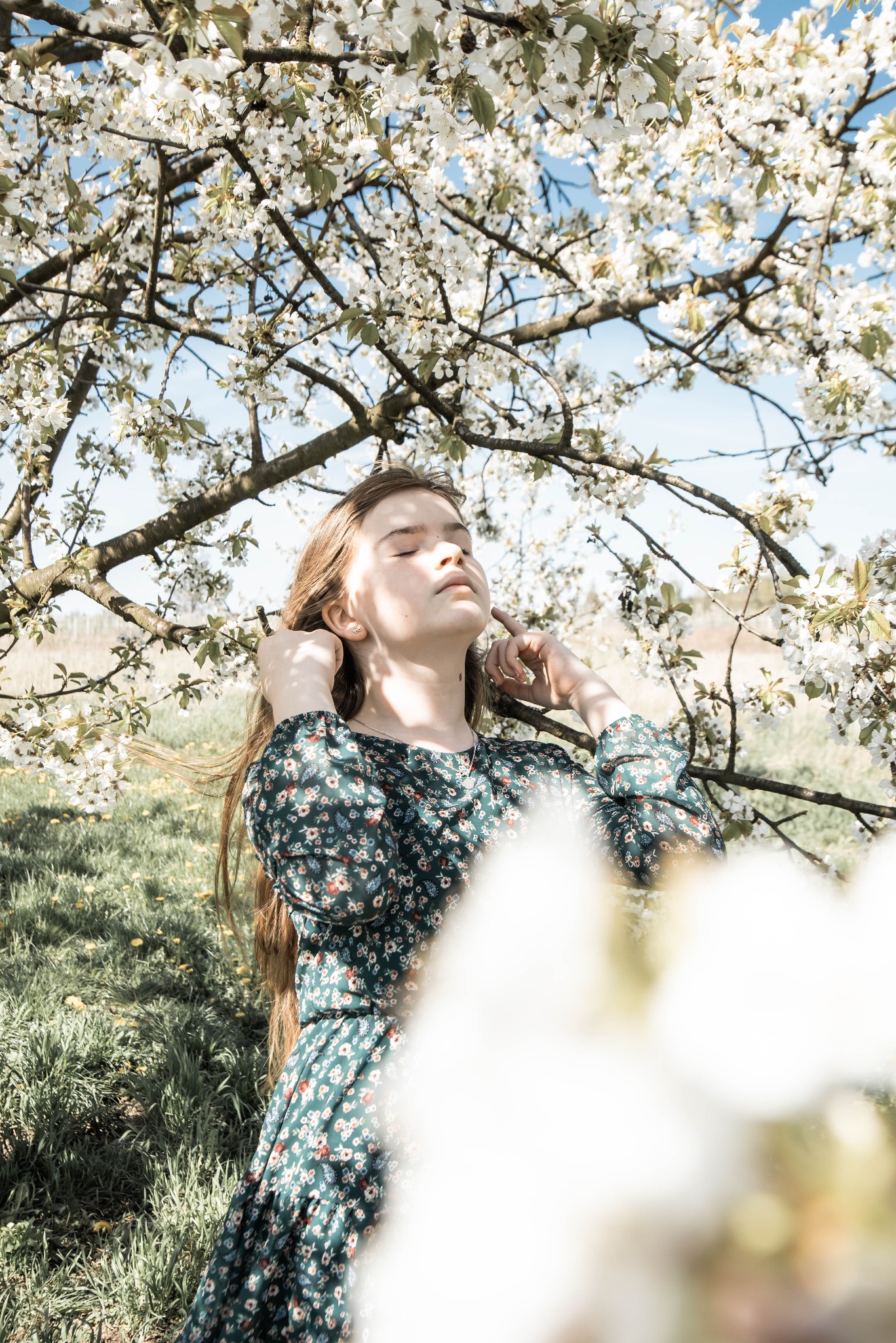 Młoda kobieta w kwiecistej sukience pozuje pod kwitnącym drzewem, dotykając delikatnie włosów, z zamkniętymi oczami w słoneczny dzień.