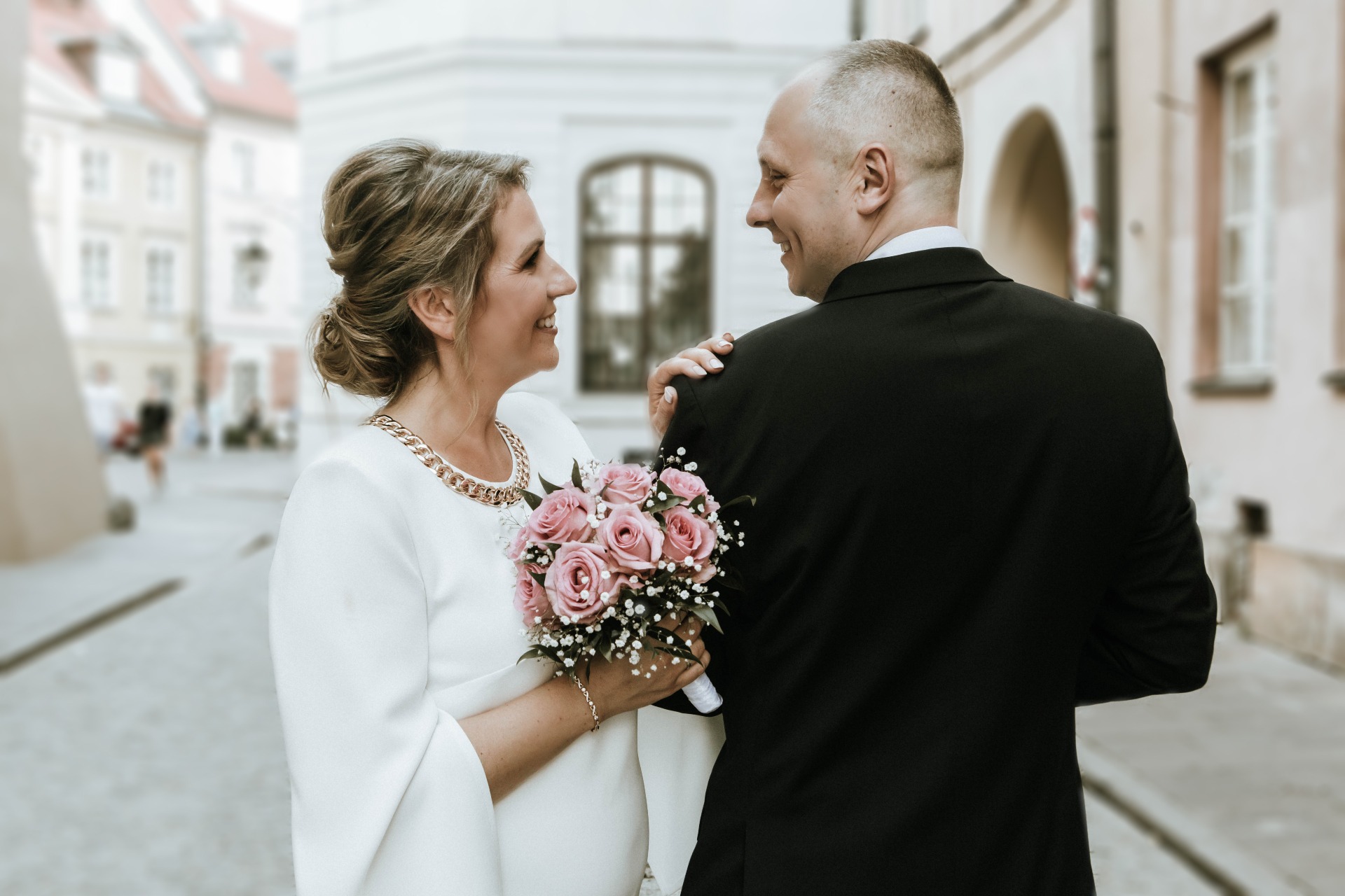 Uśmiechnięta para młoda, panna młoda w białej sukience z bukietem różowych róż, pan młody w czarnym garniturze, patrzą na siebie na tle miejskiej uliczki z budynkami w tle.
