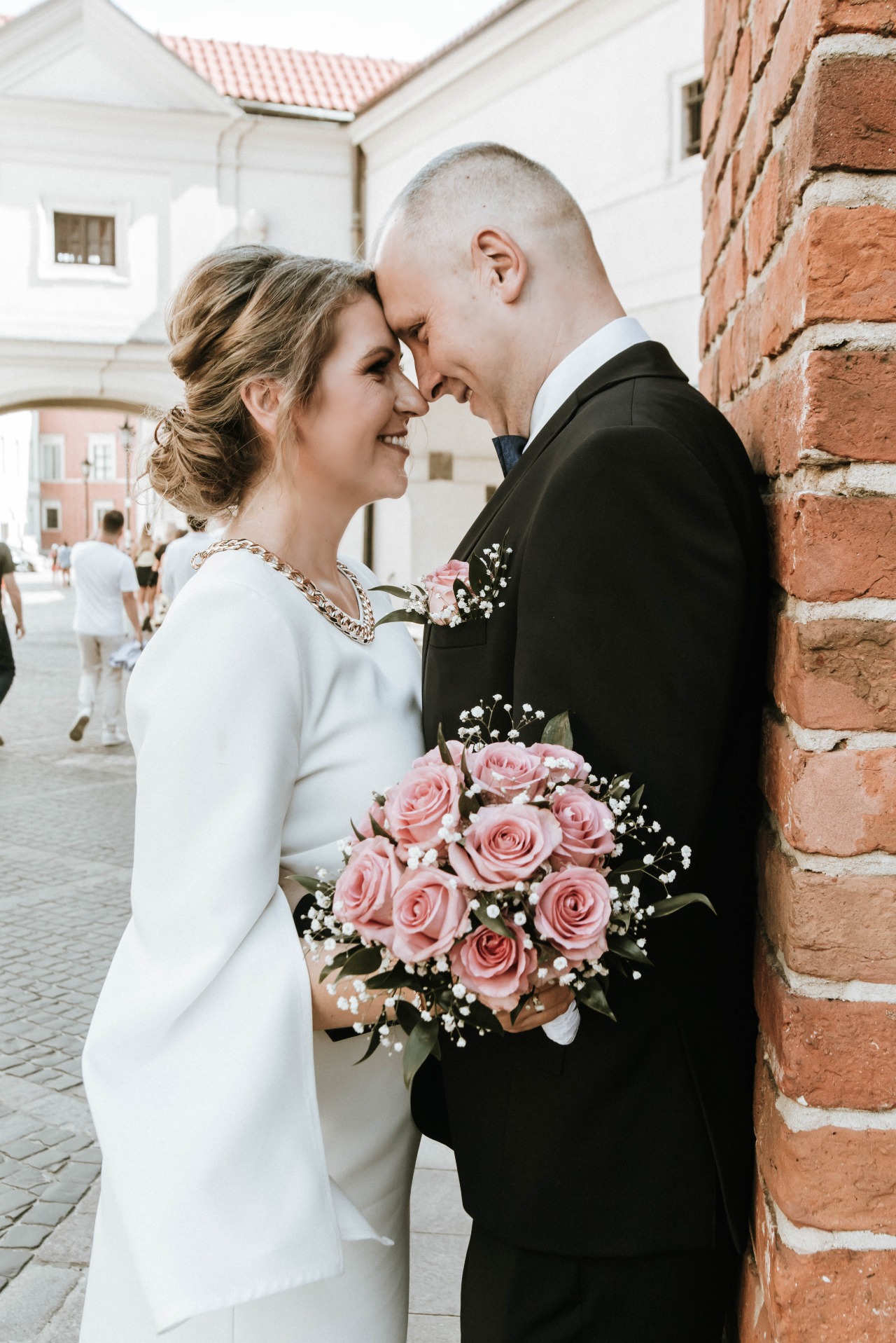 Para młoda styka się czołami, uśmiechając się do siebie, panna młoda trzyma bukiet różowych róż, a pan młody stoi przy ceglanej ścianie w czarnym garniturze z niebieską muszką.