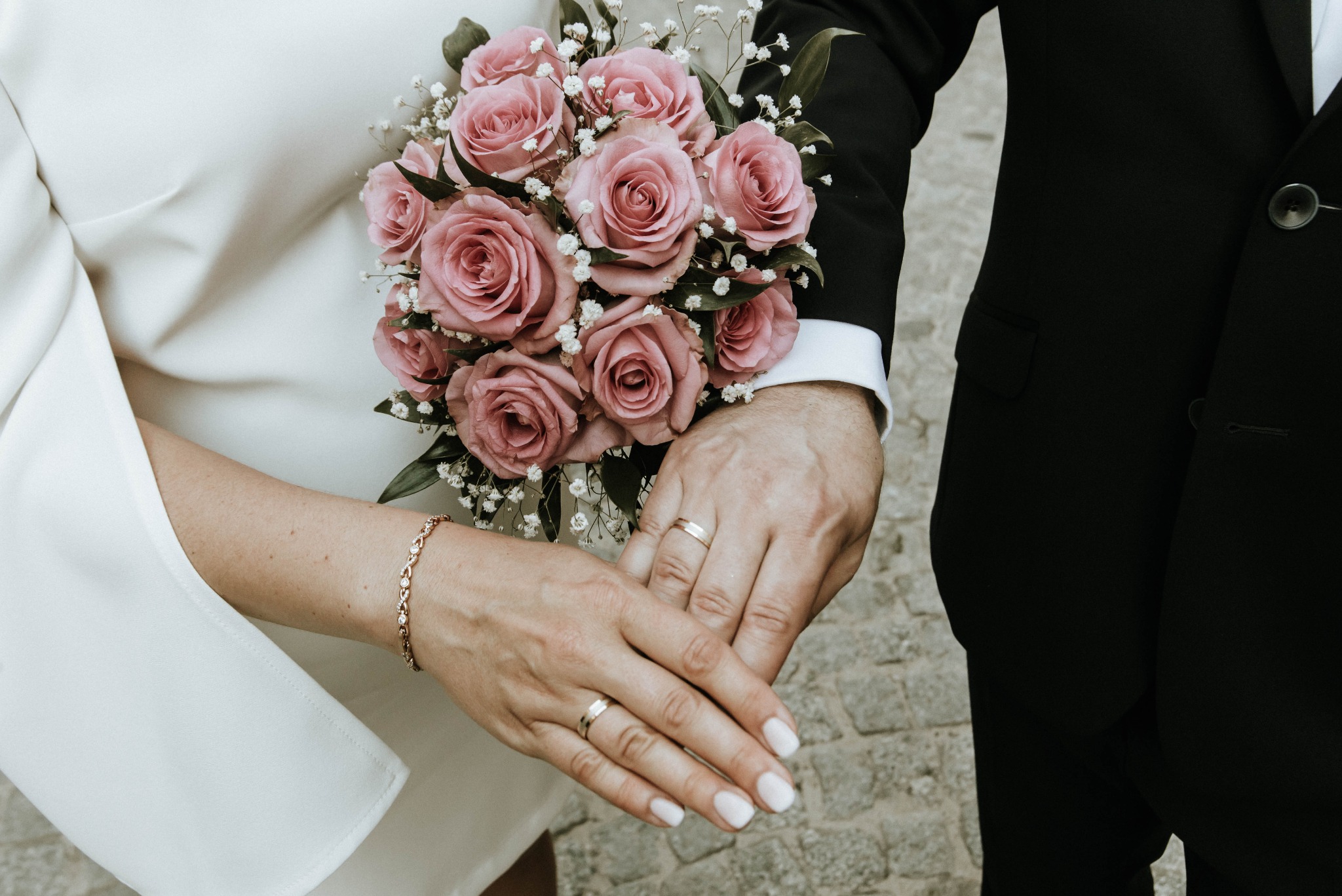 Dłonie pary młodej z obrączkami spoczywają na bukiecie z pudrowo-różowych róż i gipsówki, widoczny złoty bransoletka panny młodej i czarny garnitur pana młodego.