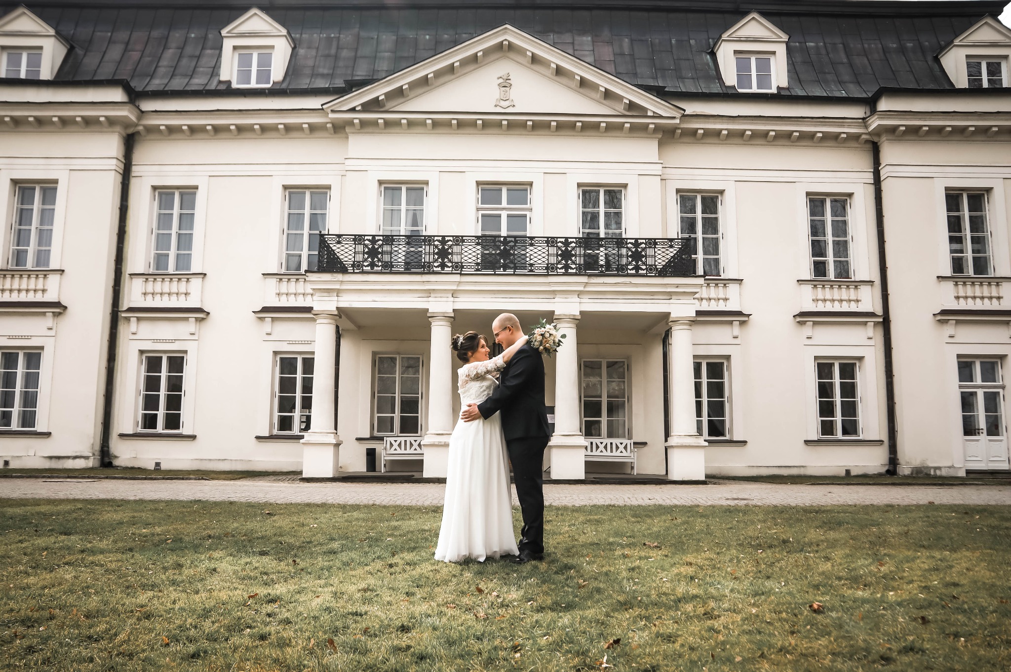 Para młoda obejmująca się na tle klasycystycznego pałacu z kolumnami i balkonem z kutą balustradą, panna młoda w sukni z koronkowymi rękawami trzyma bukiet.