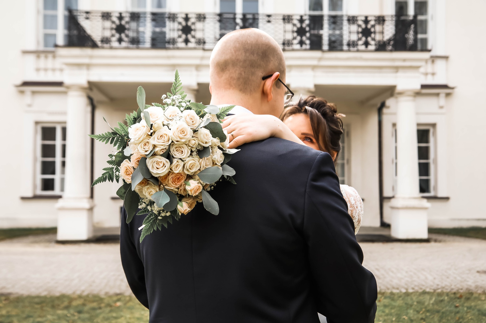Para młoda obejmująca się tyłem, bukiet z kremowych róż na plecach mężczyzny, fragment twarzy kobiety z widocznym okiem, biały budynek z balkonem w tle.