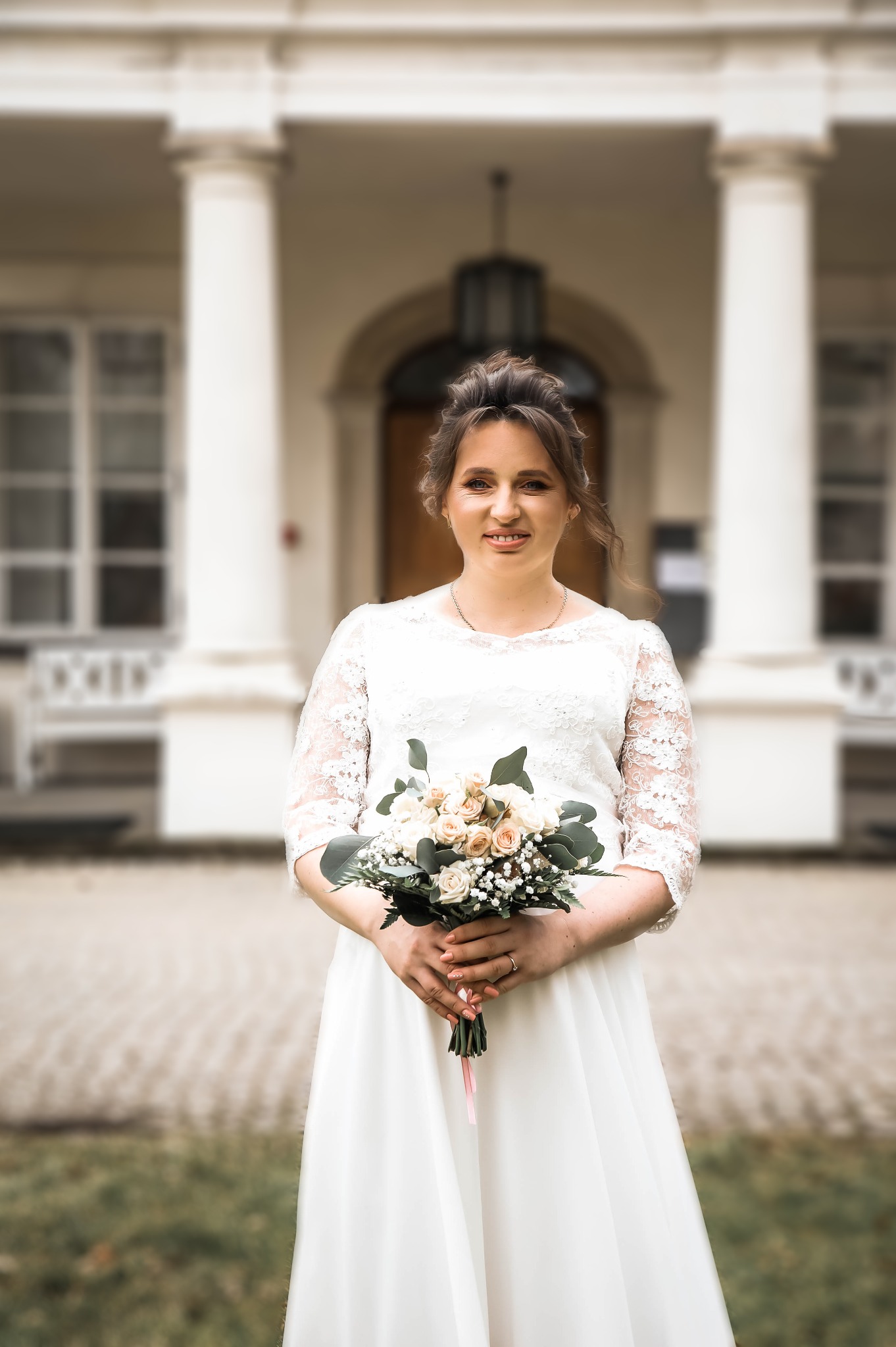 Uśmiechnięta panna młoda w koronkowej sukni trzyma bukiet z różowych i kremowych róż na tle klasycystycznego budynku z kolumnami.