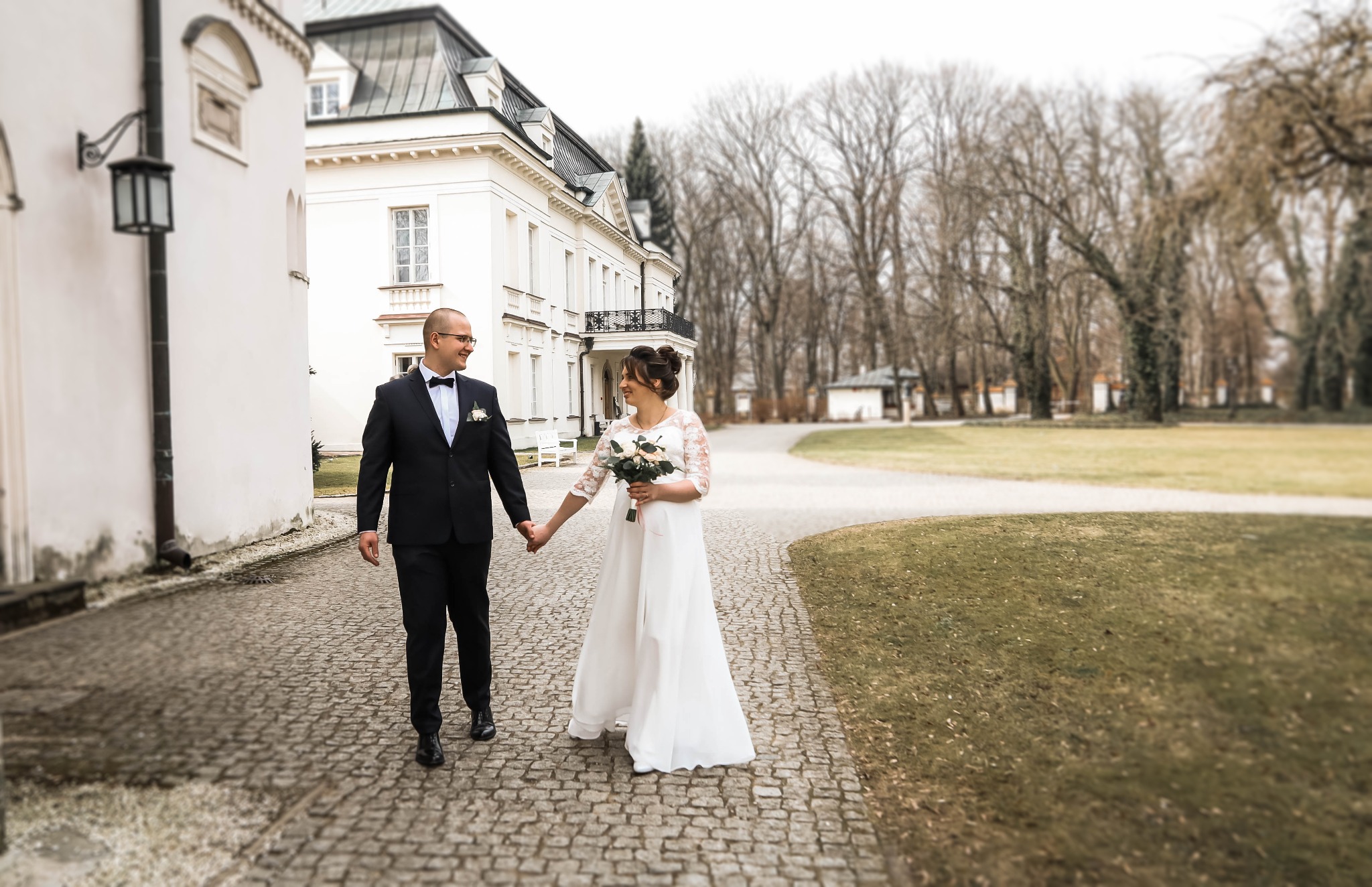 Para młoda trzymająca się za ręce spaceruje po brukowanej ścieżce przed klasycystycznym budynkiem z ciemnym dachem i parkiem w tle.