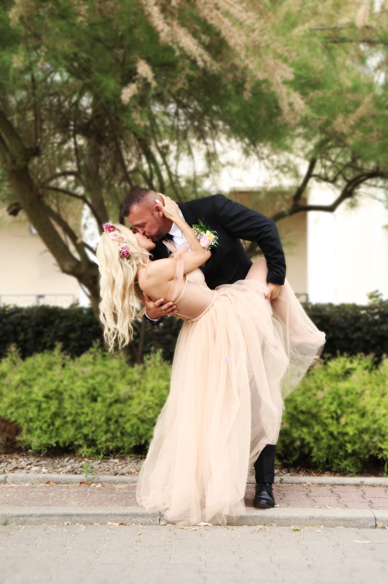 Para młoda w tańcu, panna młoda w jasnej sukni i wianku z kwiatów, pan młody w czarnym garniturze, pocałunek na tle zieleni.