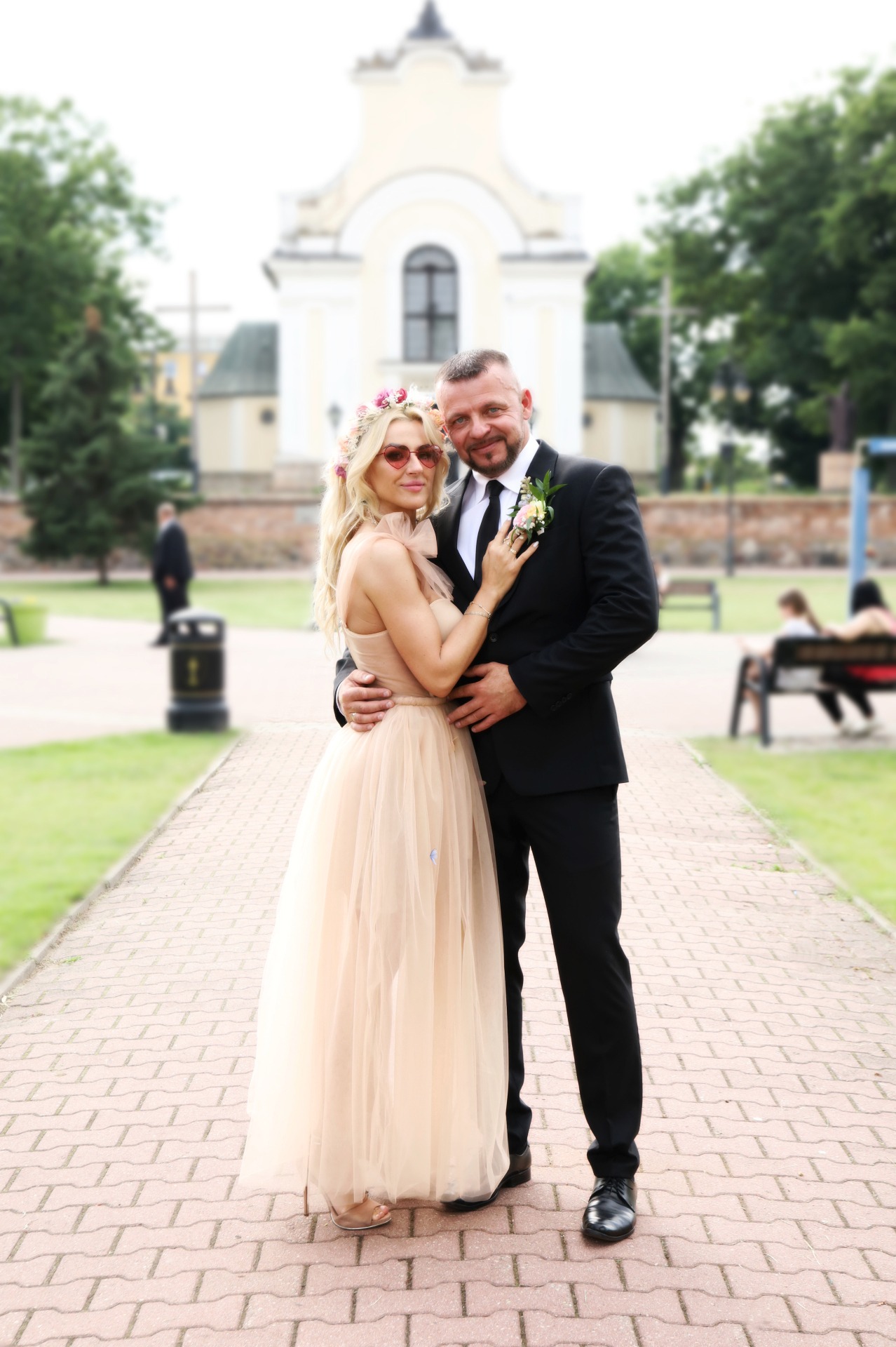 Para w strojach ślubnych pozuje na ceglanym chodniku przed kościołem w słoneczny dzień, panna młoda w okularach w kształcie serc i wianku na głowie.