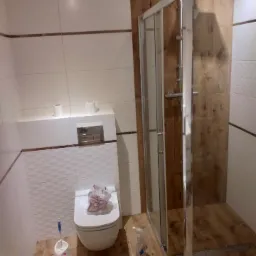 Nowoczesna łazienka z kabiną prysznicową typu walk-in, białe płytki z dekoracyjnym wzorem i drewnopodobna podłoga, widoczny wiszący sedes i akcesoria łazienkowe.