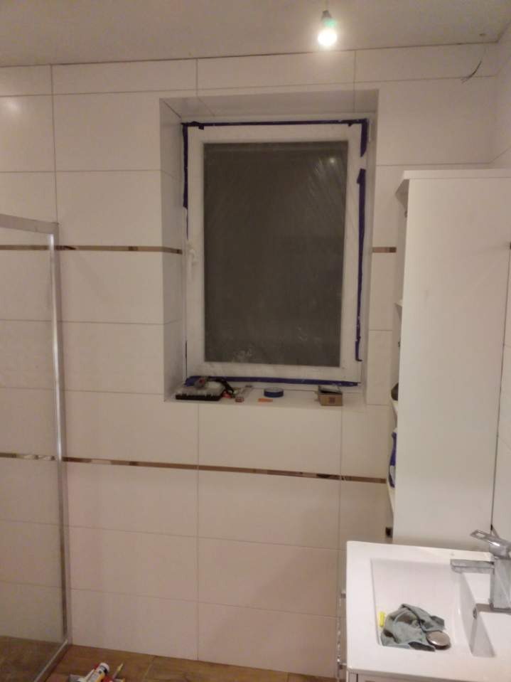 Białe płytki z metalowymi listwami w łazience, okno zabezpieczone folią, narzędzia na parapecie, umywalka z baterią i szmatką.
