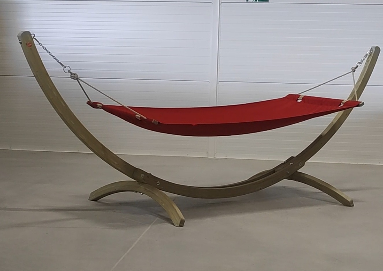 Czerwony hamak z płótna rozpięty na drewnianym stelażu, widok z boku na tle białej ściany z paneli.