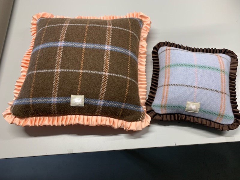 Dwie kwadratowe poduszki dekoracyjne w kratę z falbankami: większa brązowa z pomarańczową falbanką i mniejsza błękitna z brązową falbanką, obie z małą białą naszywką w rogu.