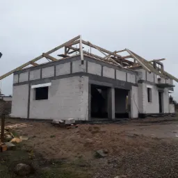 Surowy stan budowy: ściany z betonu komórkowego parterowego domu z widoczną konstrukcją drewnianą dachu, teren wokół w stanie roboczym, pochmurne niebo.