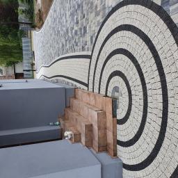 Dam-Bud - Ułożona kostka brukowa z dekoracyjnym wzorem koncentrycznych okręgów, prowadząca do schodów wejściowych budynku. W tle widoczny fragment posesji z zielenią.