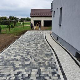 Dam-Bud - Ułożona z szaro-białej kostki brukowej ścieżka prowadząca do budynku. Dwóch mężczyzn z żółtym wężem w tle, trawnik z huśtawką po lewej stronie i szary budynek po prawej.