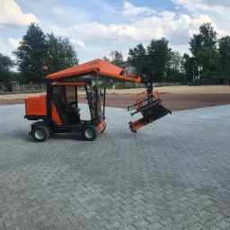 Pomarańczowy manipulator do kostki brukowej Optimas H 88 na tle ułożonej szarej kostki, w tle przygotowany teren i drzewa.