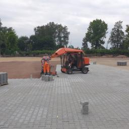 Dam-Bud - Pracownik w pomarańczowym kombinezonie układa szare kostki brukowe przy użyciu specjalistycznej maszyny do układania kostki; paleta z kostką brukową w tle.