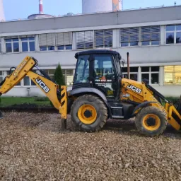 Żółta koparko-ładowarka JCB na żwirowym podłożu, w tle budynek przemysłowy z kominem, sugerująca przygotowanie terenu pod budowę lub układanie kostki.