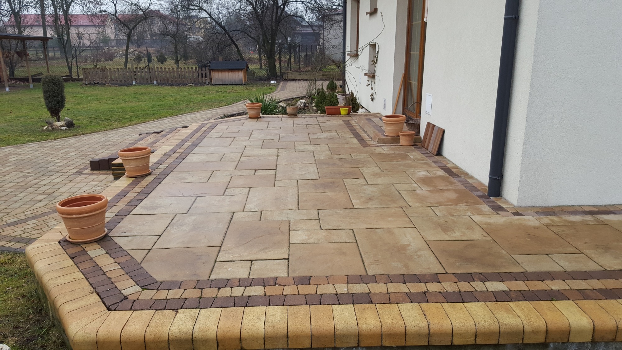 Wykończony taras z kostki brukowej w odcieniach beżu i brązu, obrzeża z ciemniejszej kostki, ozdobne donice z terakoty, w tle trawnik i zabudowania.