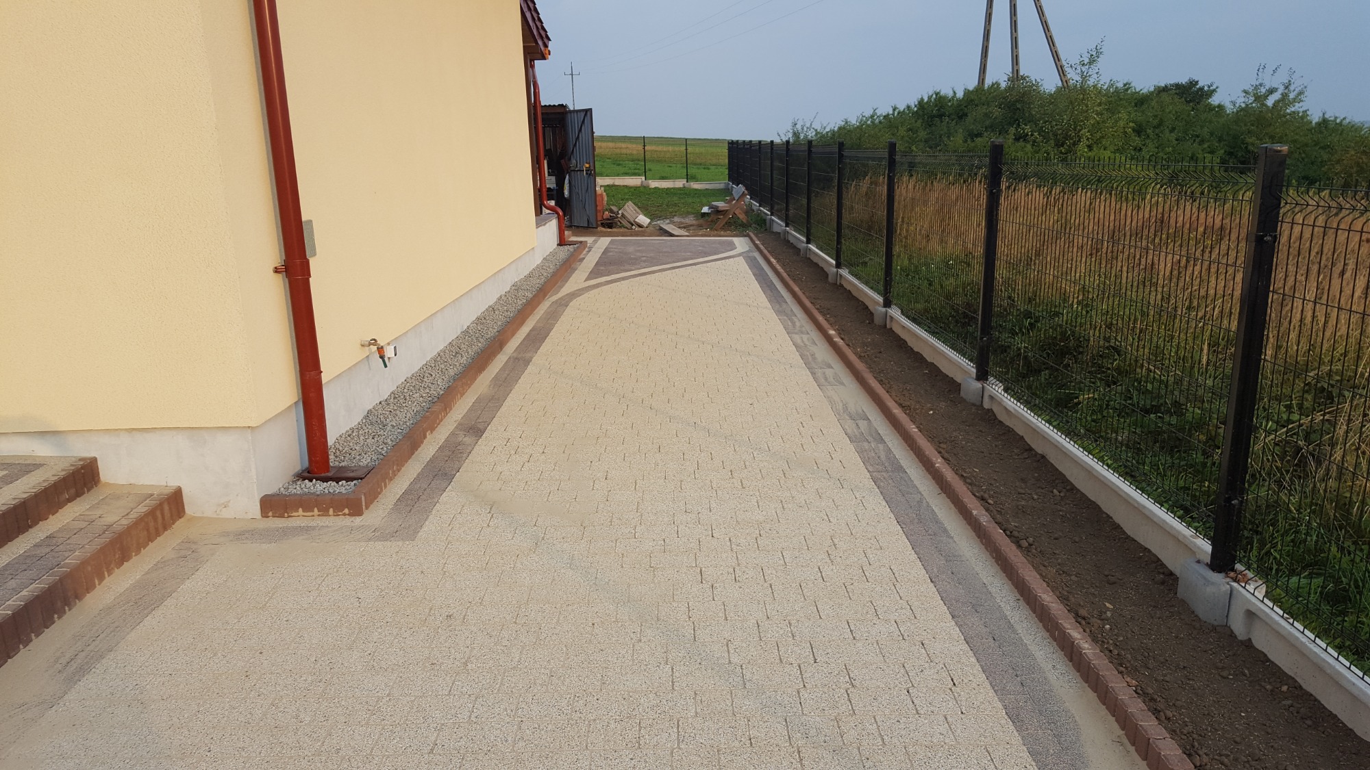 Ułożona kostka brukowa wzdłuż domu i ogrodzenia panelowego, z widocznymi schodami wejściowymi i fragmentem trawnika za ogrodzeniem.