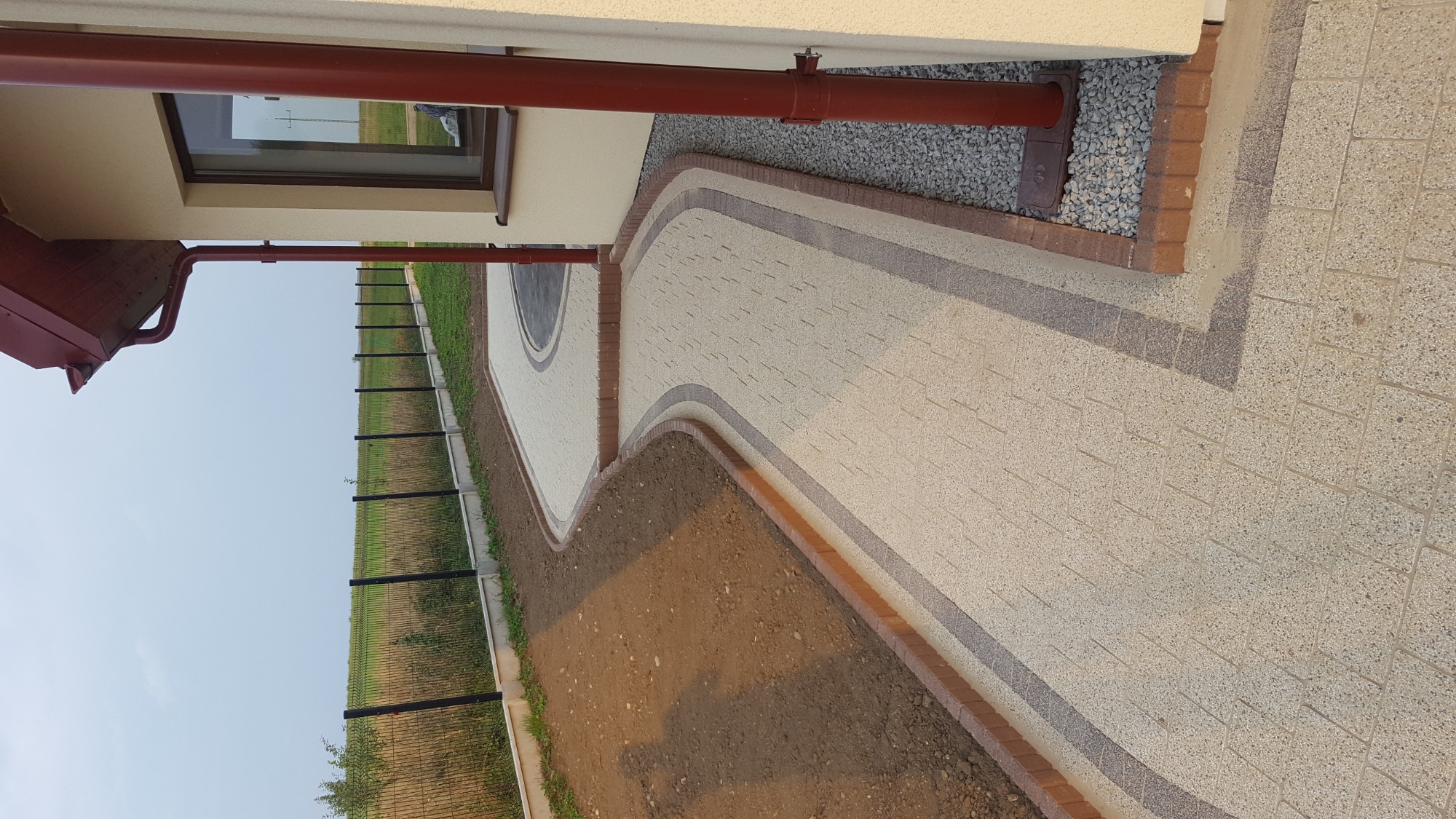 Ułożona ścieżka z kostki brukowej w odcieniach beżu i szarości, z obrzeżami z czerwonej cegły, prowadząca wzdłuż domu z rynną i oknem, obok przygotowany teren pod trawnik i ogrodzenie panelowe.